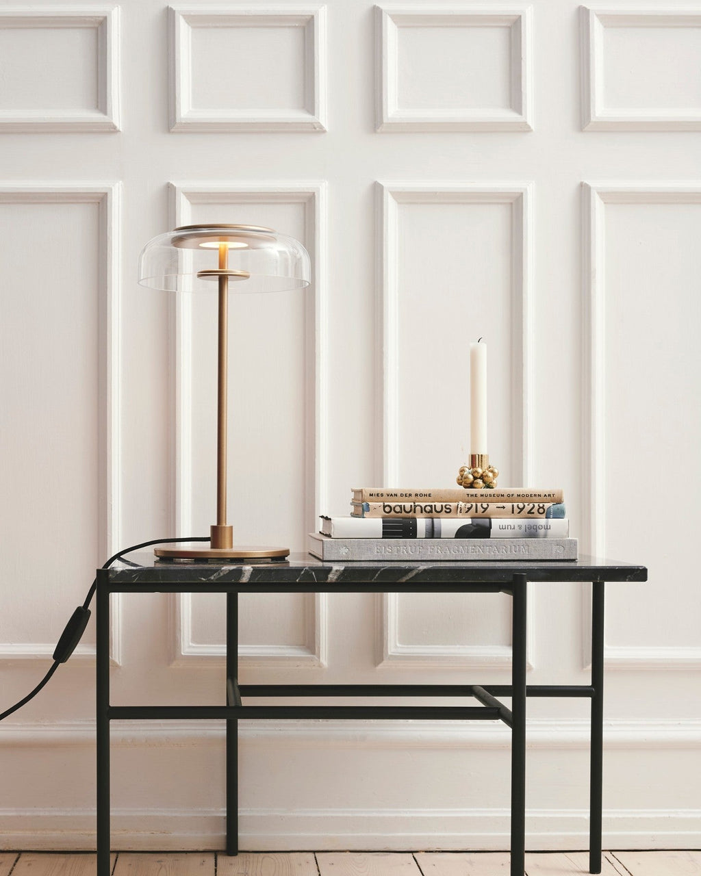 A Clérachic table lamp lit on a marble-topped black table, with a taper candle on a stack of books, against white wall panels.