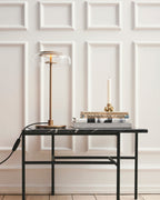 A Clérachic table lamp lit on a marble-topped black table, with a taper candle on a stack of books, against white wall panels.