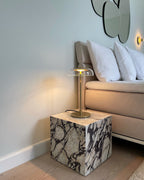 A lit Clérachic table lamp on a marble cube beside a sofa with white cushions, under a pair of round mirrors on a pale wall.