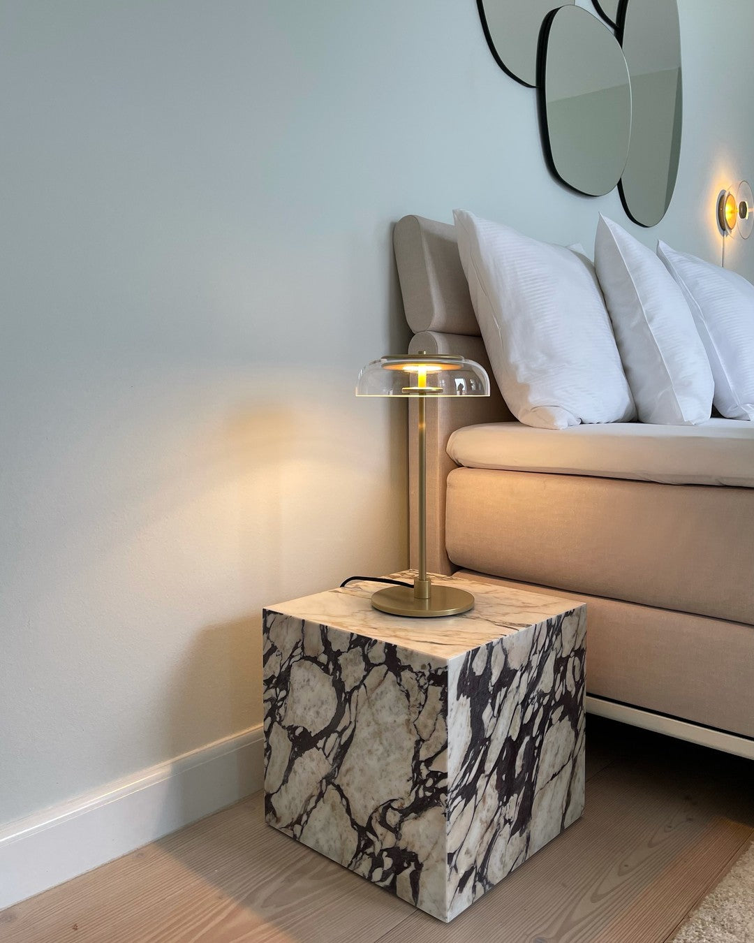 A lit Clérachic table lamp on a marble cube beside a sofa with white cushions, under a pair of round mirrors on a pale wall.