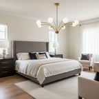 Above a bed with a grey upholstered headboard, a Clérachic chandelier with white globes on slim brass arms in a bright bedroom.
