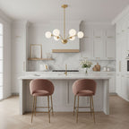 A Clérachic brass chandelier with white globes above a marble-topped kitchen island, with two pink velvet bar stools in front.