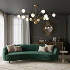 A Clérachic chandelier with white globes above a curved green velvet sofa, with a sunburst mirror on a dark wall behind.