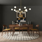 Above a round walnut dining table with mustard chairs, a Clérachic brass chandelier with white globes against a dark grey wall.