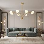 A Clérachic brass chandelier with white globes in a living room with a sage-green sofa, a brass coffee table and tall wall uplights.