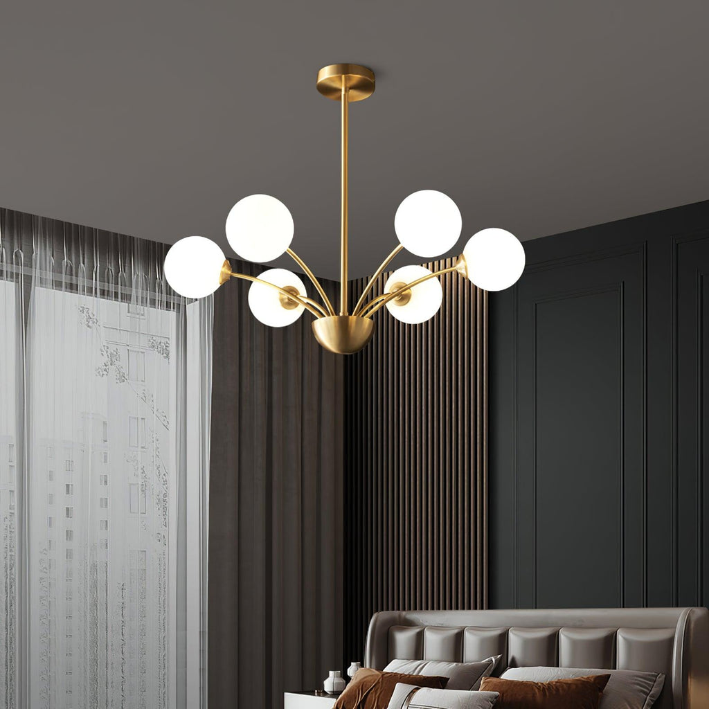 A Clérachic brass chandelier with white globes in a bedroom with grey panelled walls, sheer curtains and a tufted headboard below.