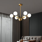 A Clérachic brass chandelier with white globes in a bedroom with grey panelled walls, sheer curtains and a tufted headboard below.
