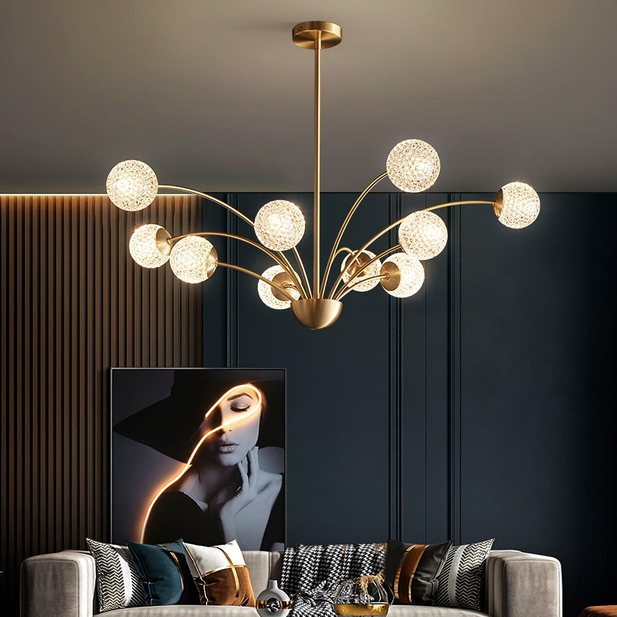A Clérachic brass chandelier with warm textured globes over a grey sofa, with a framed portrait on a dark panelled wall.