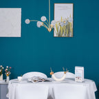 A Clérachic chandelier with textured globes in an unlit state against a bright teal wall, with botanical posters and a white-clothed table below.