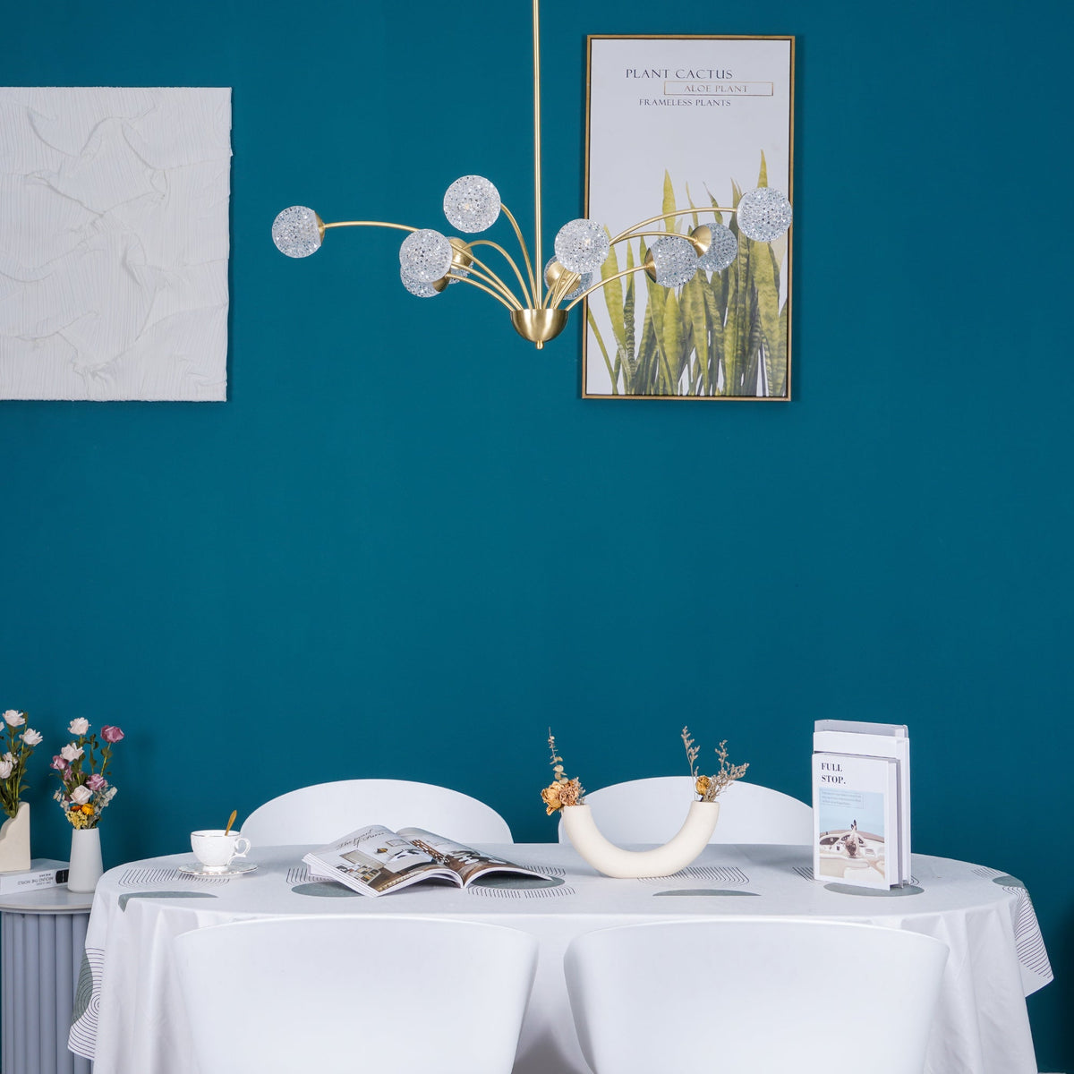 A Clérachic chandelier with textured globes in an unlit state against a bright teal wall, with botanical posters and a white-clothed table below.