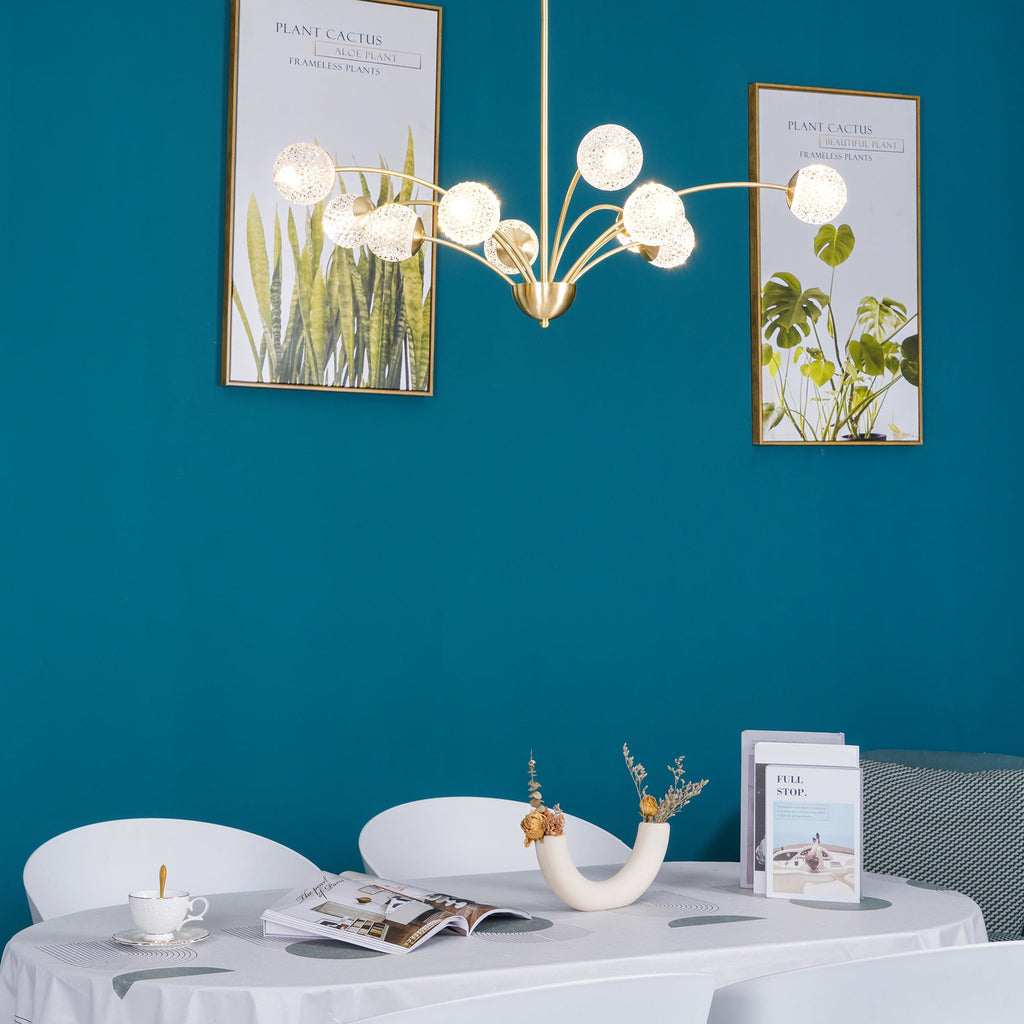 A Clérachic chandelier with its textured globes glowing, set against a teal wall with two framed plant prints above a dining table.