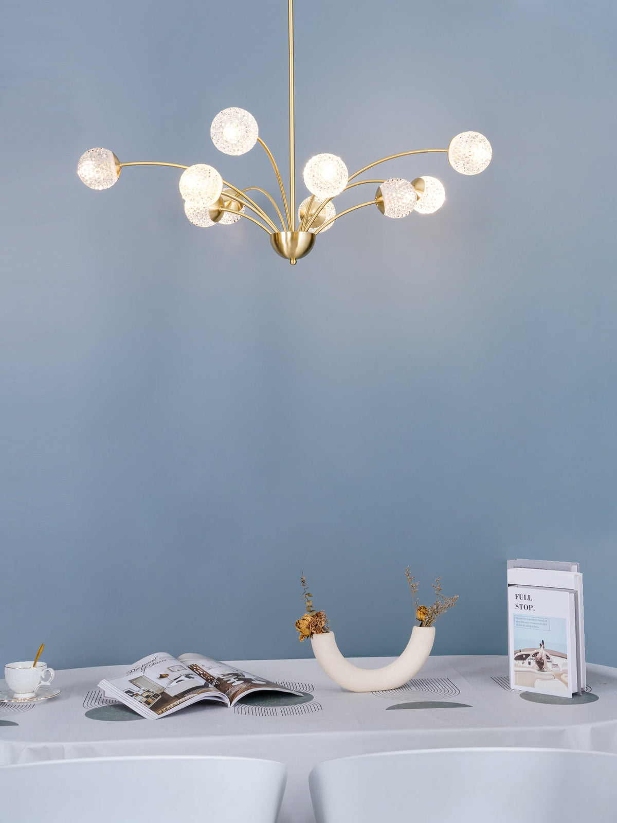 A Clérachic brass chandelier with textured globes lit against a pale blue wall, with a white-clothed table and a vase below.