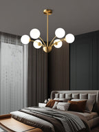 A Clérachic brass chandelier with white globes above a dark bedroom with a grey upholstered bed and layered cushions.