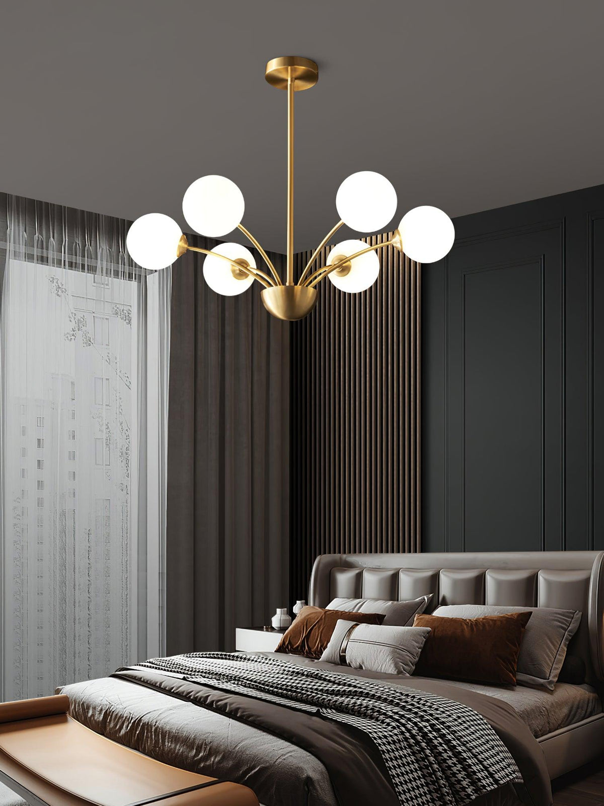 A Clérachic brass chandelier with white globes above a dark bedroom with a grey upholstered bed and layered cushions.