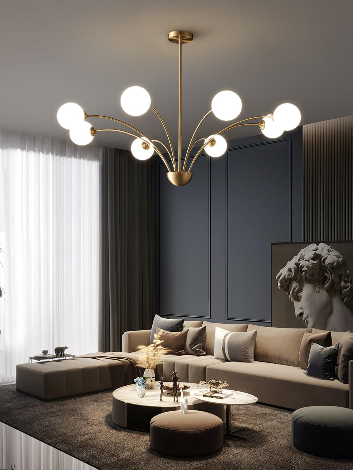 A Clérachic brass chandelier with white globes over a beige sectional sofa, with a classical bust and sheer curtains behind.