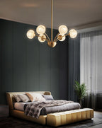 A Clérachic brass chandelier with textured globes lit over a gold-edged bed, in a dark green bedroom with a tall window.
