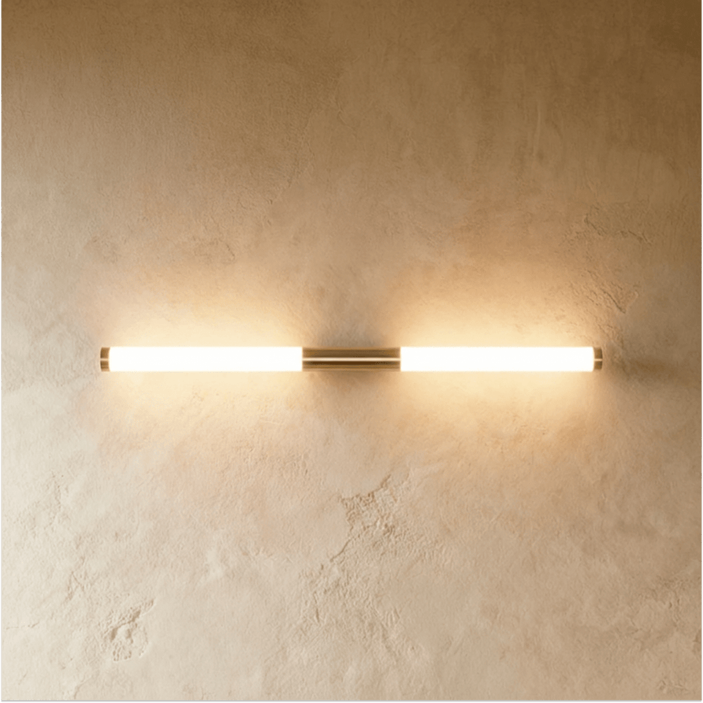 Clérachic slim brass-finish LED tube wall light glowing warm on a textured plaster wall, mounted horizontally.