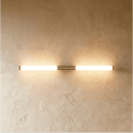 Clérachic slim brass-finish LED tube wall light glowing warm on a textured plaster wall, mounted horizontally.