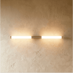 Clérachic slim brass-finish LED tube wall light glowing warm on a textured plaster wall, mounted horizontally.