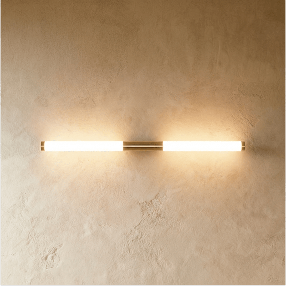 Clérachic slim brass-finish LED tube wall light glowing warm on a textured plaster wall, mounted horizontally.