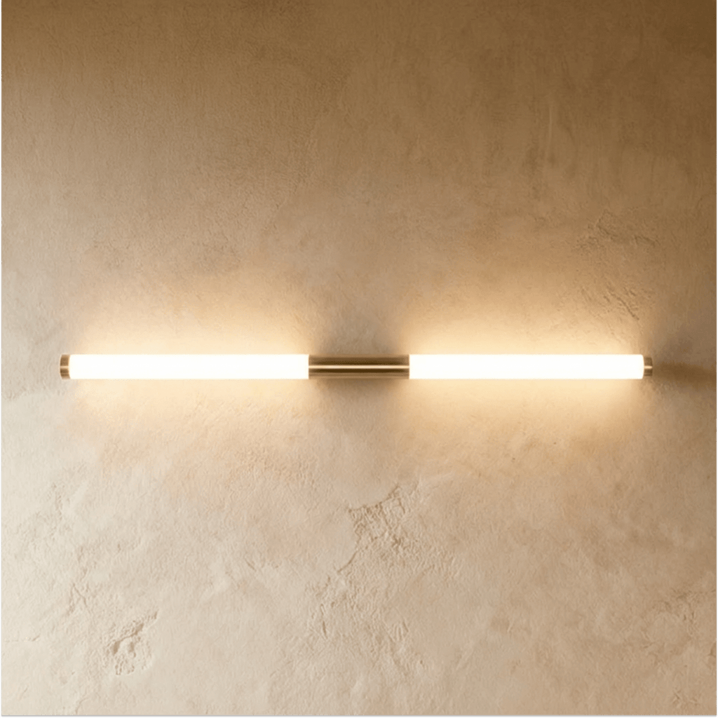 Clérachic slim brass-finish wall light with two frosted glowing tubes and brass end caps on a textured plaster wall.