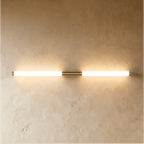 Clérachic slim brass-finish wall light with two frosted glowing tubes and brass end caps on a textured plaster wall.