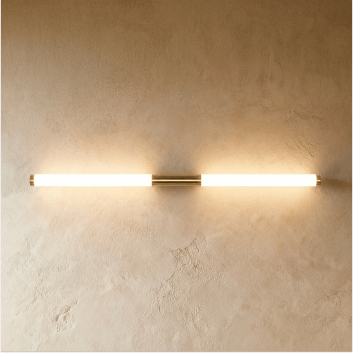 Clérachic slim brass-finish wall light with two frosted glowing tubes and brass end caps on a textured plaster wall.