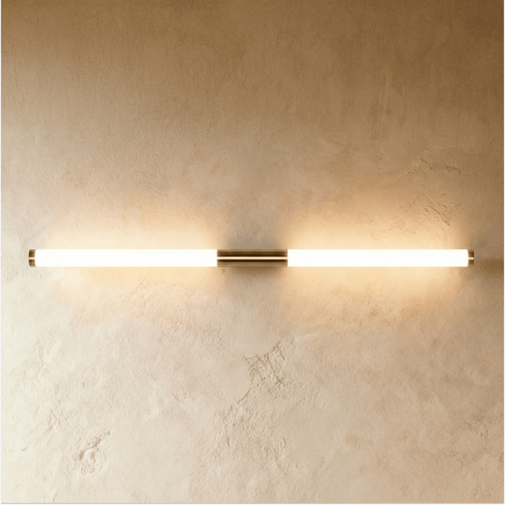 Clérachic long linear wall light glowing warm, with brass end caps and a centre mount on a pale textured wall.