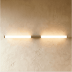 Clérachic long linear wall light glowing warm, with brass end caps and a centre mount on a pale textured wall.