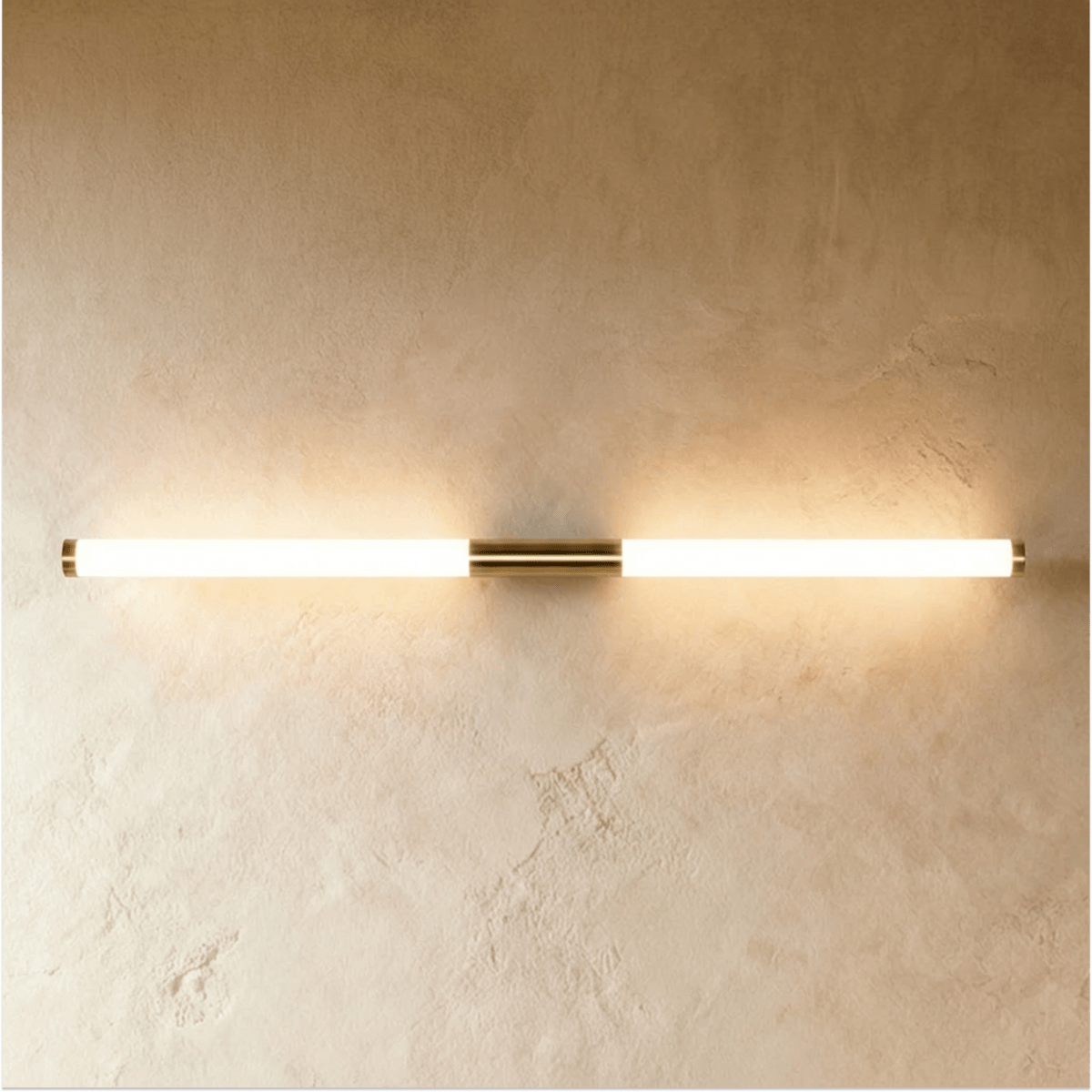 Clérachic long linear wall light glowing warm, with brass end caps and a centre mount on a pale textured wall.