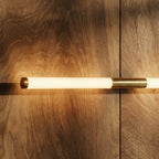 Close view of a Clérachic frosted tube wall light with a brass end cap and brass centre mount, lit on a wood-panel wall.