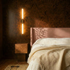 Clérachic slim vertical wall light glowing warm on burr-wood panelling beside a pink upholstered bed and cream throw.