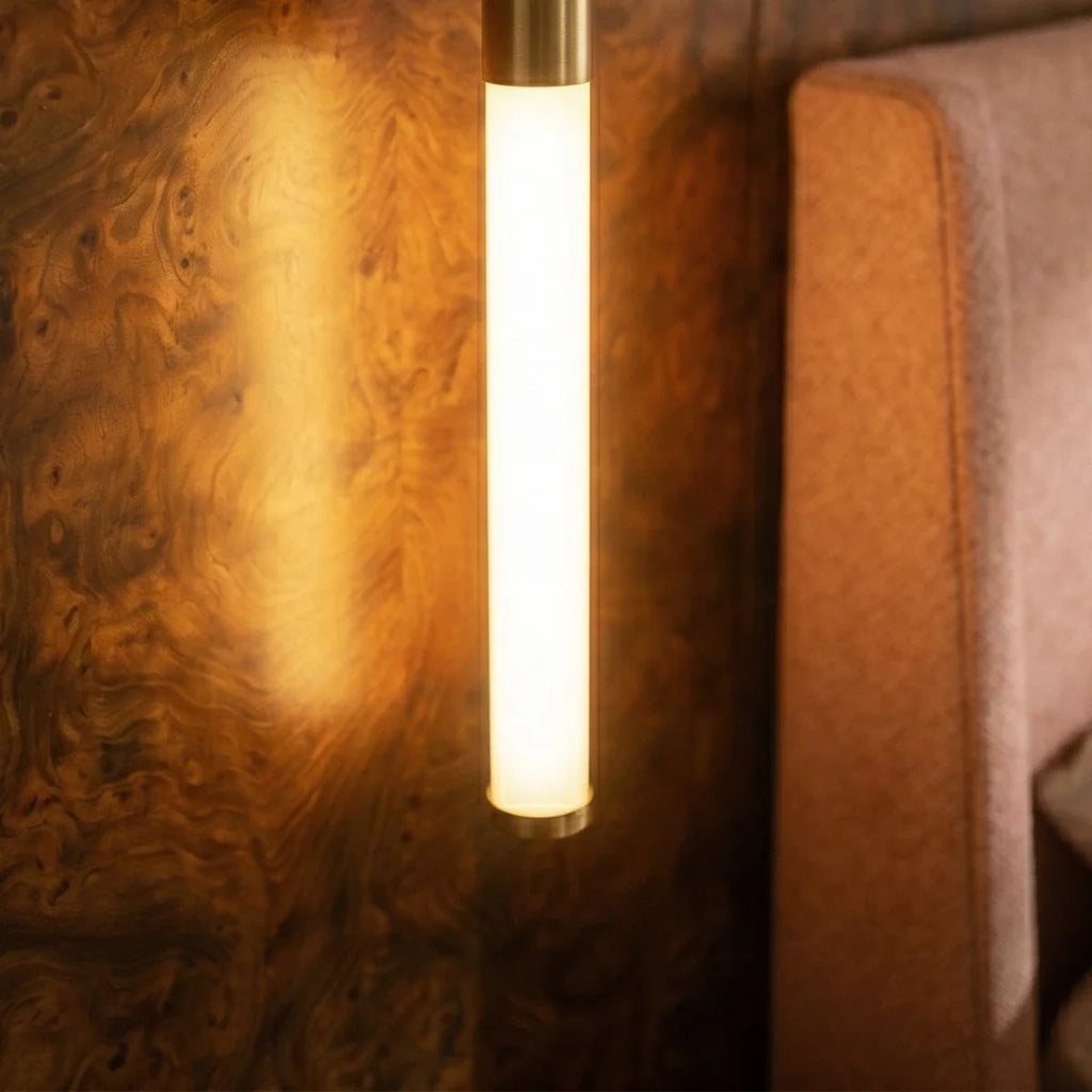 Close view of the Clérachic frosted LED tube with a brass cap, lit warm against burr-wood panelling and a pink headboard.