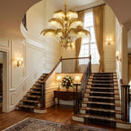 Above a sweeping staircase with a patterned runner, a Clérachic brass and crystal chandelier lights the cream hallway.