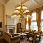 Gold brass leaves glow warmly on this Clérachic chandelier, hung in a double-height living room with gold curtains and a stone fireplace.