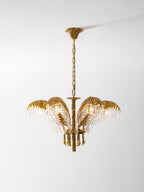 A small Clérachic brass chandelier with crystal-fringed leaves, hanging from a ceiling plate in front of a light backdrop.