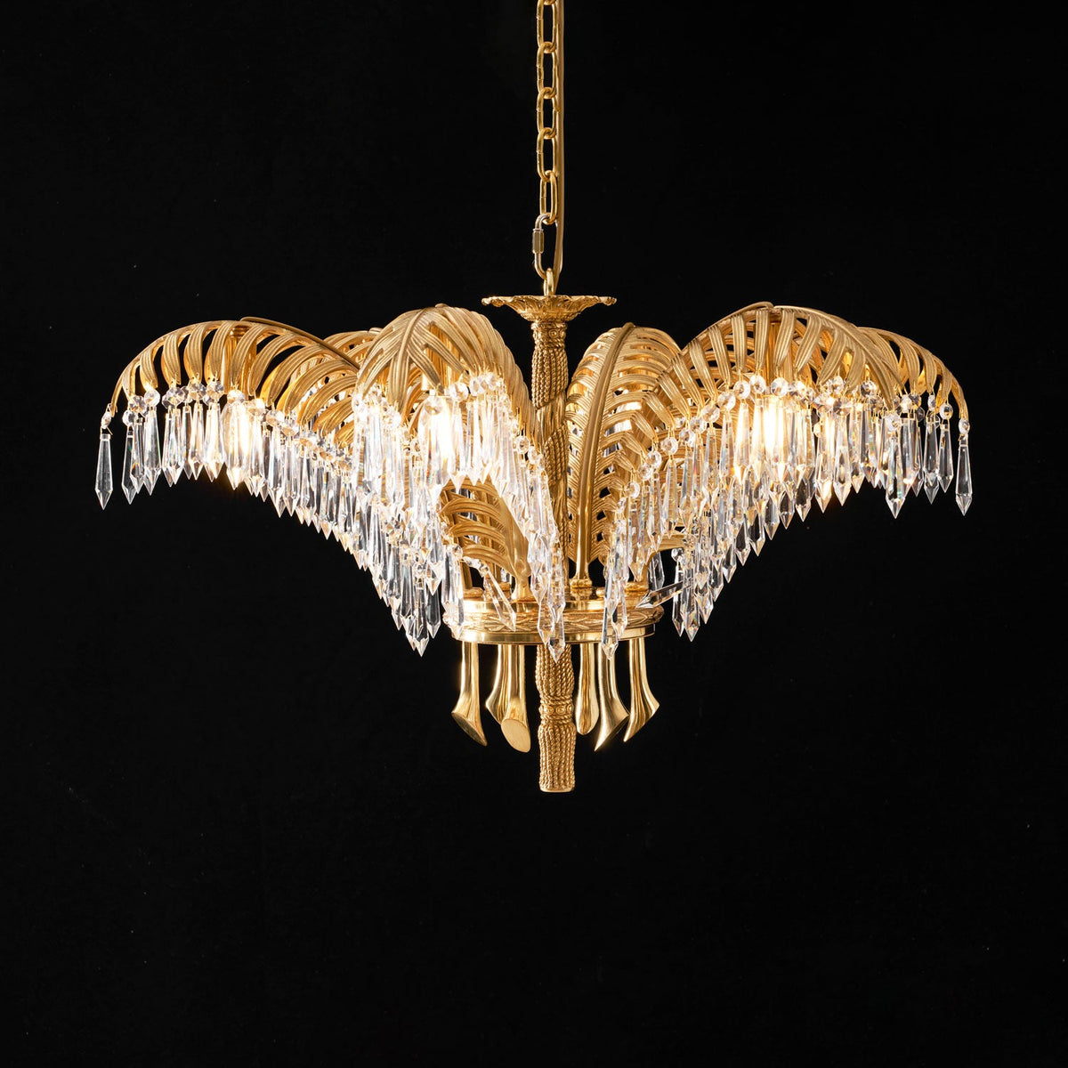 A Clérachic chandelier in lit brass and clear crystal, hung on a gold chain against a black backdrop.