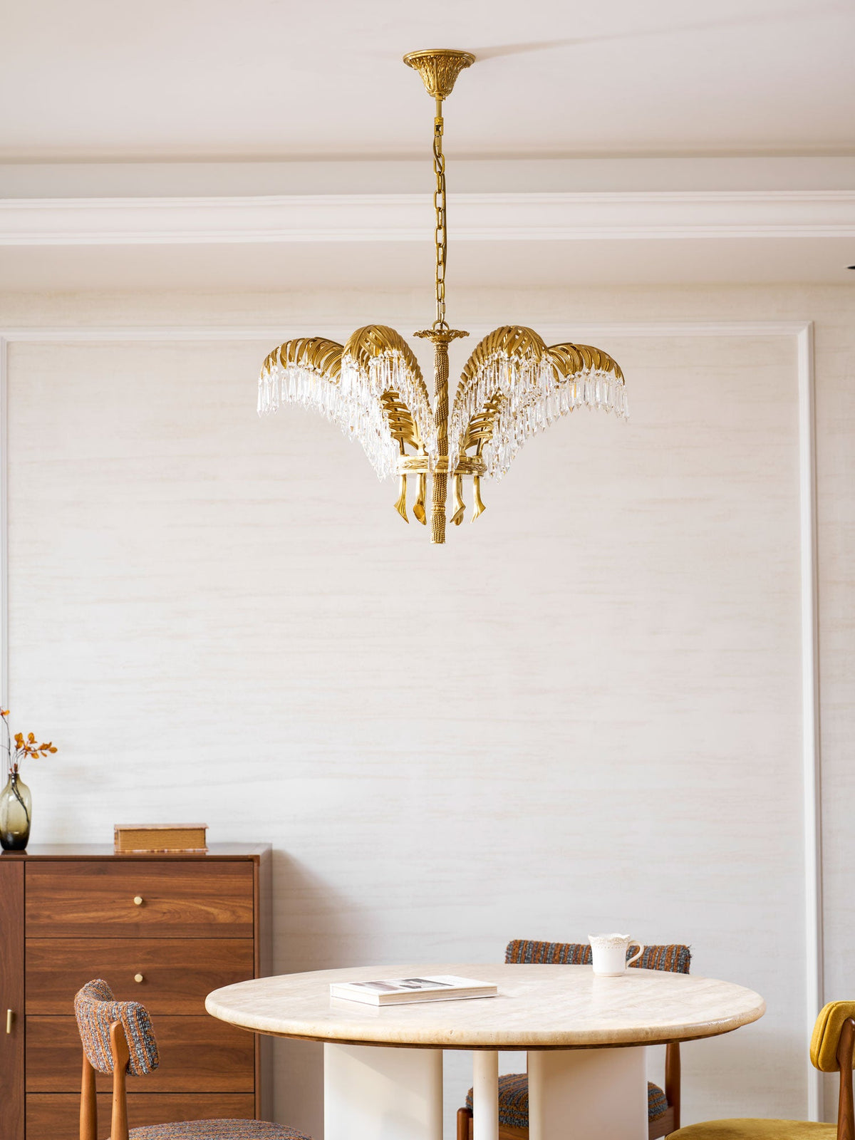 A Clérachic brass chandelier hangs above a round table with a book and a cup, with a sideboard and a vase to the left.