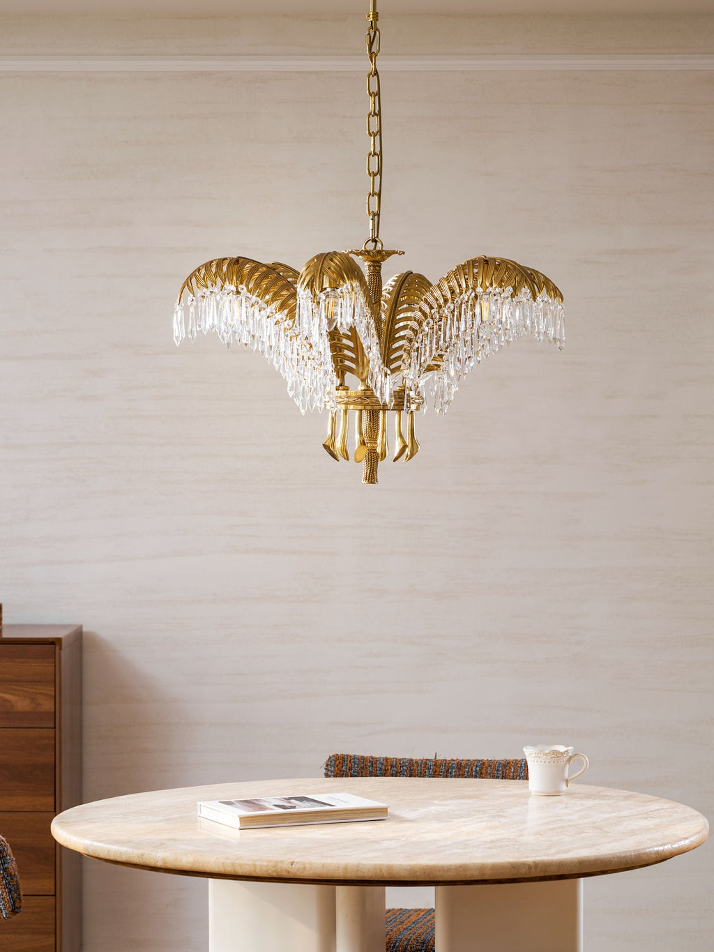Clérachic brass chandelier hung at ceiling height above a round stone-topped table, with a wooden cabinet at the left.
