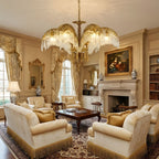 Warmly lit brass leaves of a Clérachic chandelier hang over a cream sitting room with a fireplace and a framed painting.