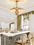Over a white-topped kitchen island with gilded detail and upholstered stools, a small Clérachic brass chandelier hangs from an ornate ceiling.