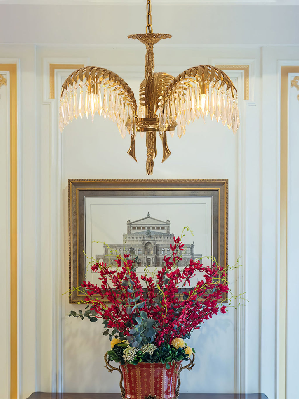 A three-arm Clérachic brass chandelier hangs in front of a framed print and a vase of red flowers in a pale hallway.