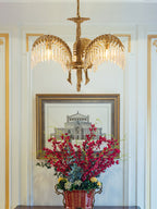 A three-arm Clérachic brass chandelier hangs in front of a framed print and a vase of red flowers in a pale hallway.