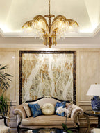 A Clérachic chandelier with brass leaves hangs in a cream room with a marble feature wall and a sofa with blue cushions.