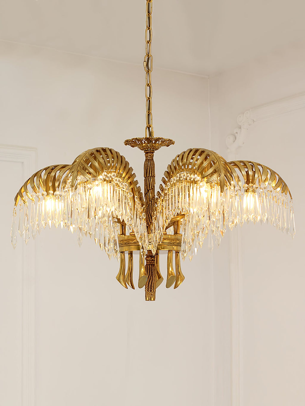 A Clérachic brass chandelier hangs against a pale wall with moulding, its leaves lit warmly from within.