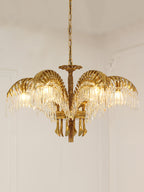 A Clérachic brass chandelier hangs against a pale wall with moulding, its leaves lit warmly from within.