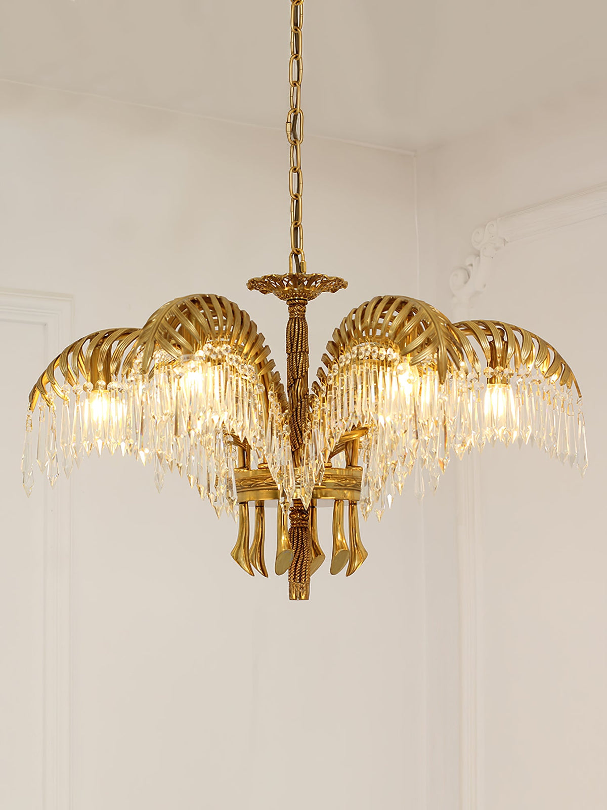 A Clérachic brass chandelier hangs against a pale wall with moulding, its leaves lit warmly from within.