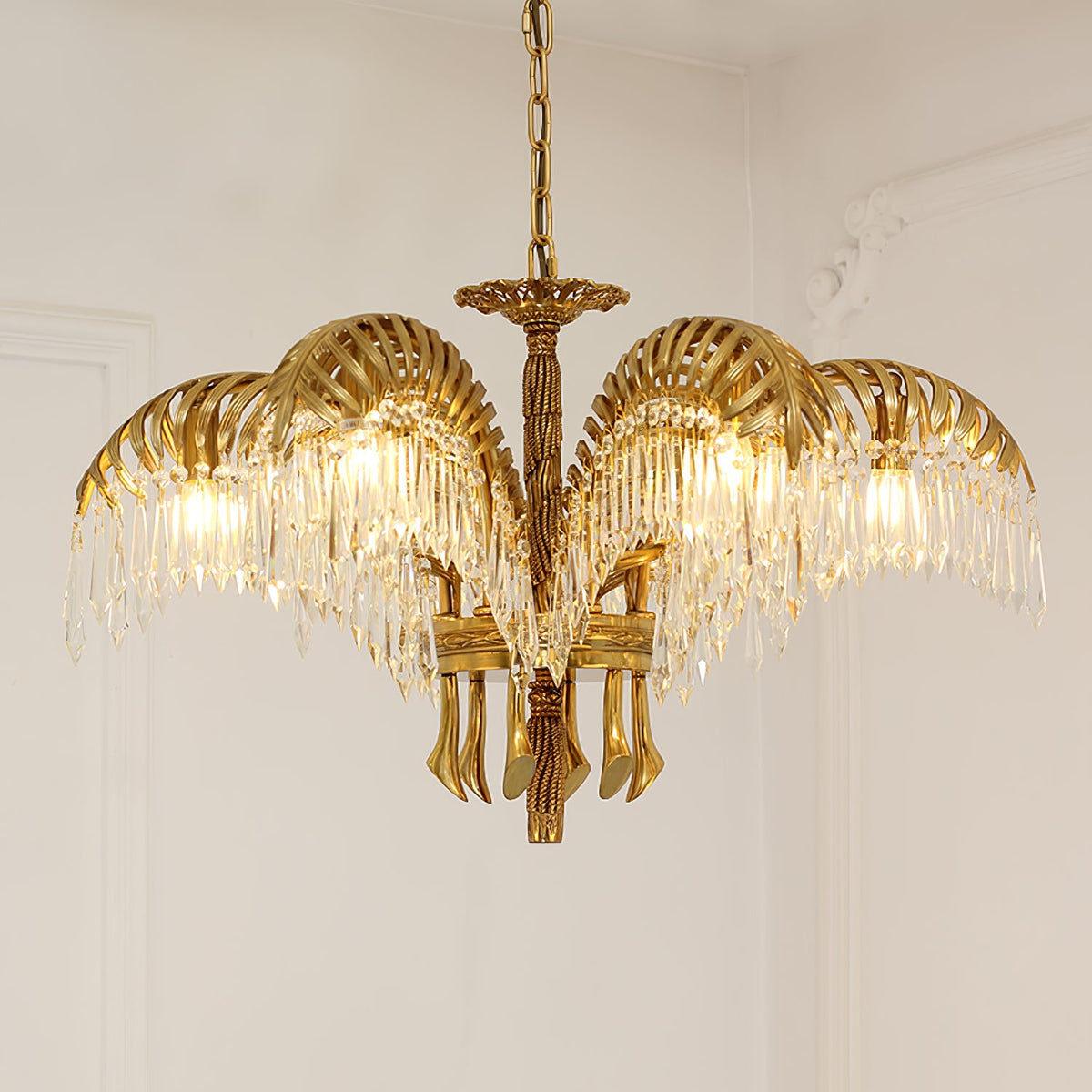 A Clérachic gold brass chandelier on a chain hangs against a pale wall with plaster moulding, its crystal fringes glowing.