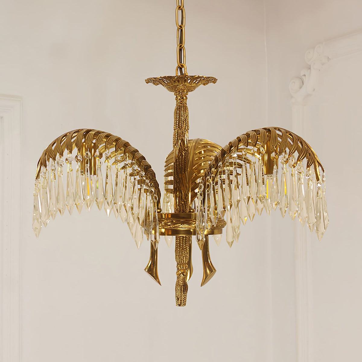 Front view of a three-leaf Clérachic chandelier in brass with crystal fringes, hanging on a chain against a pale wall.