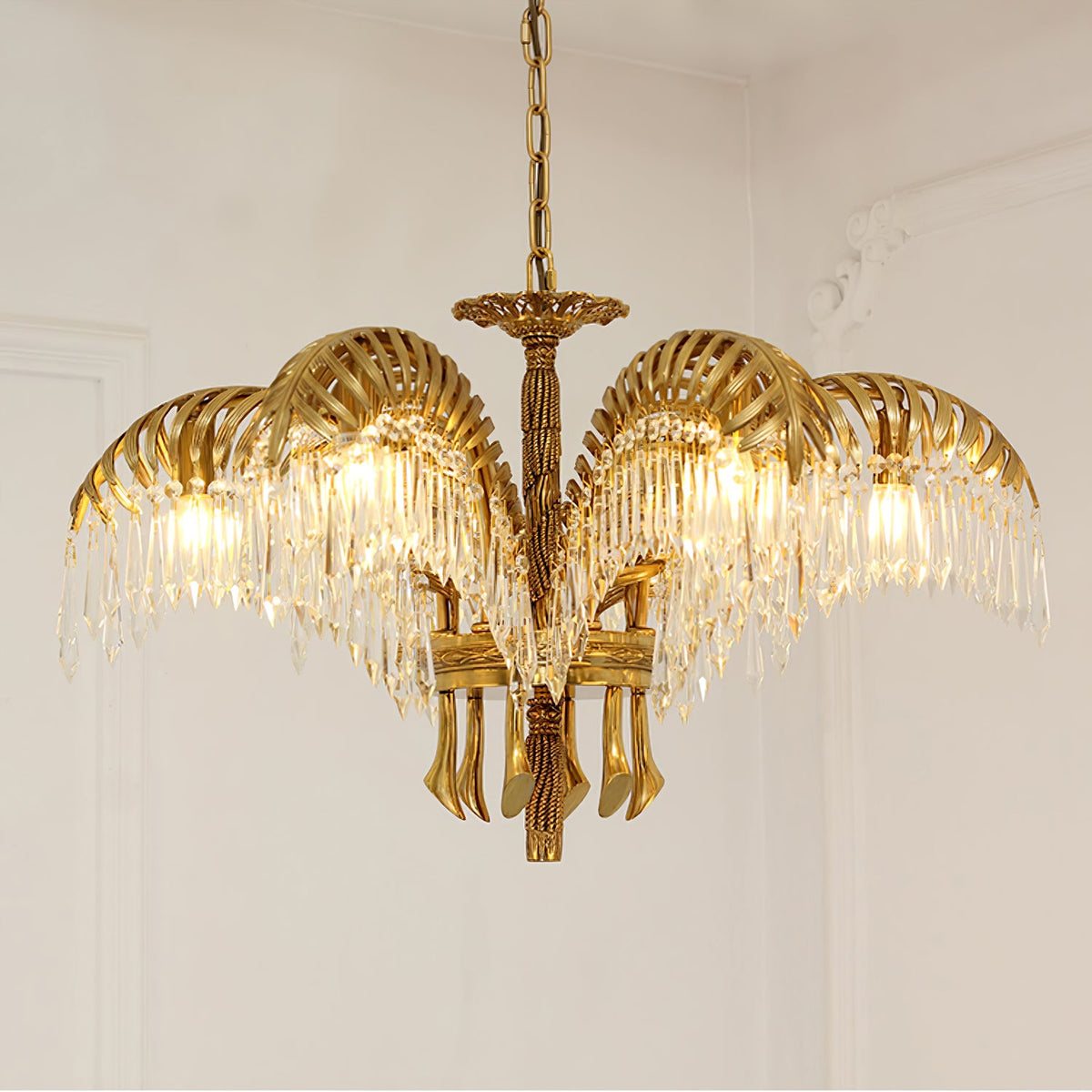 Lit from within, this Clérachic gold brass chandelier shows its crystal-fringed leaves in front of a pale moulded wall.